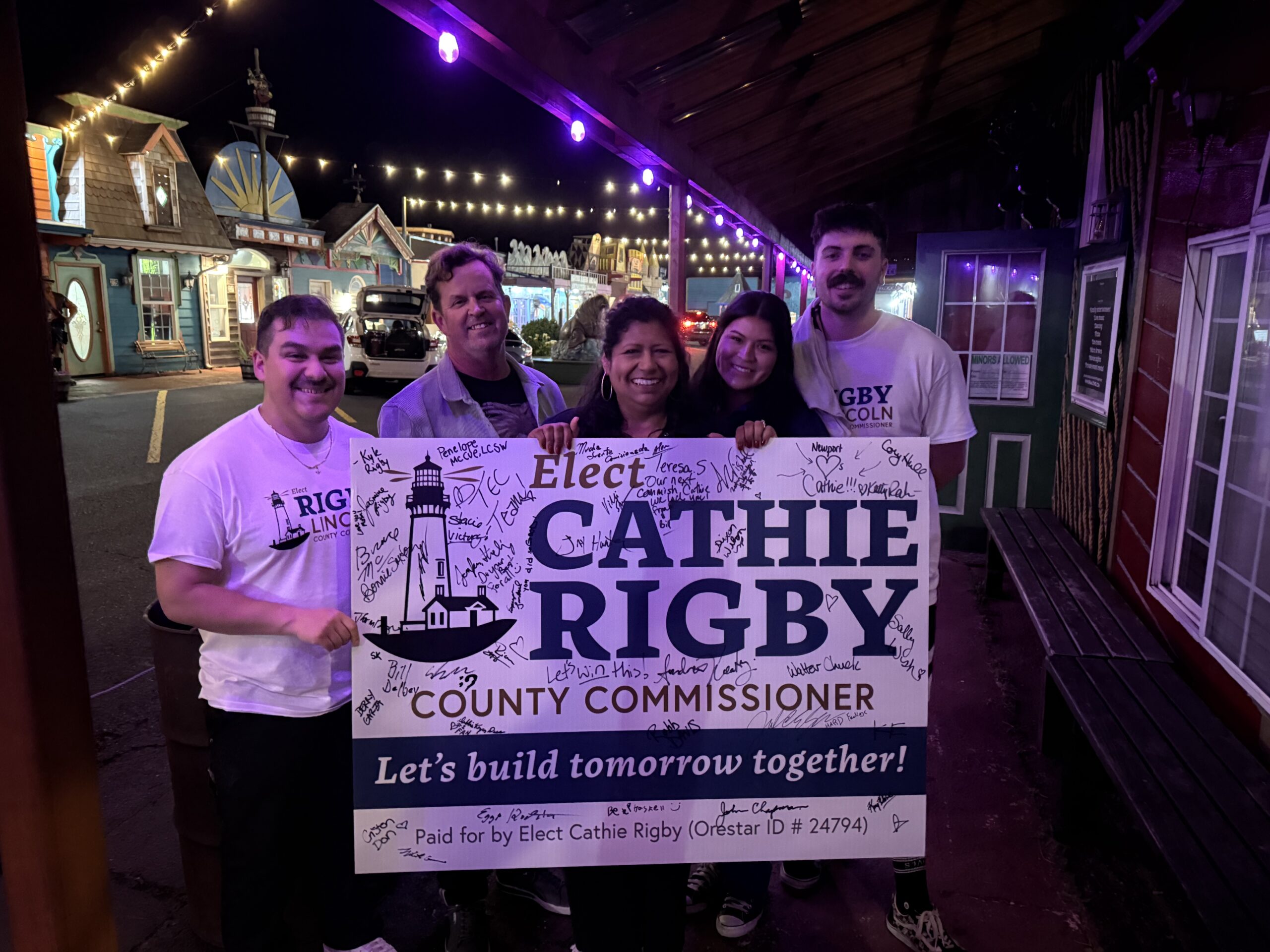 Five people pose together holding a large campaign banner reading 'Elect Cathie Rigby for County Commissioner' at night under string lights on a colorful street set.