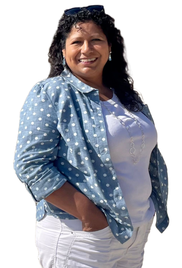 Smiling woman on a sandy beach wearing a blue polka-dot shirt over a white top and white pants, sunglasses on her head.